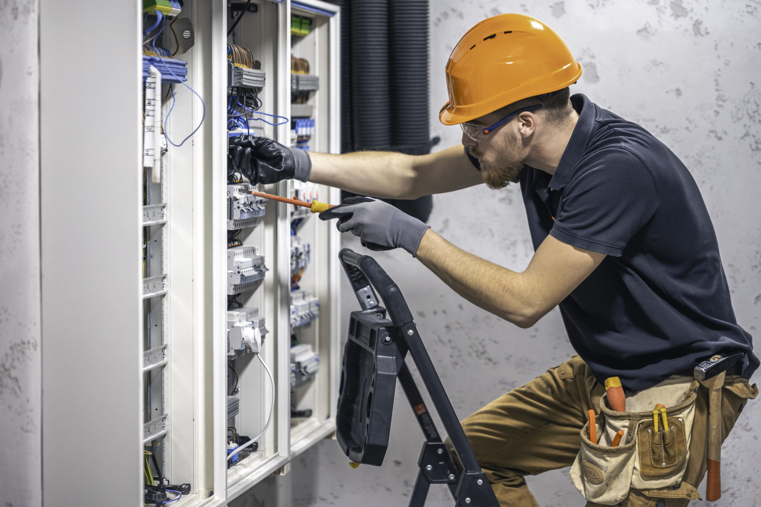 A male electrician works in a switchboard with an electrical connecting cable. Electrician with screwdriver tightens electrically operated switching equipment in fuse box.